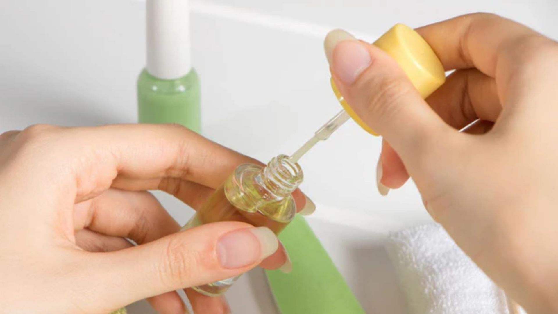 Hands holding a nail oil bottle with a dropper. The setting includes a green bottle and a white towel, conveying a serene, self-care ambiance.