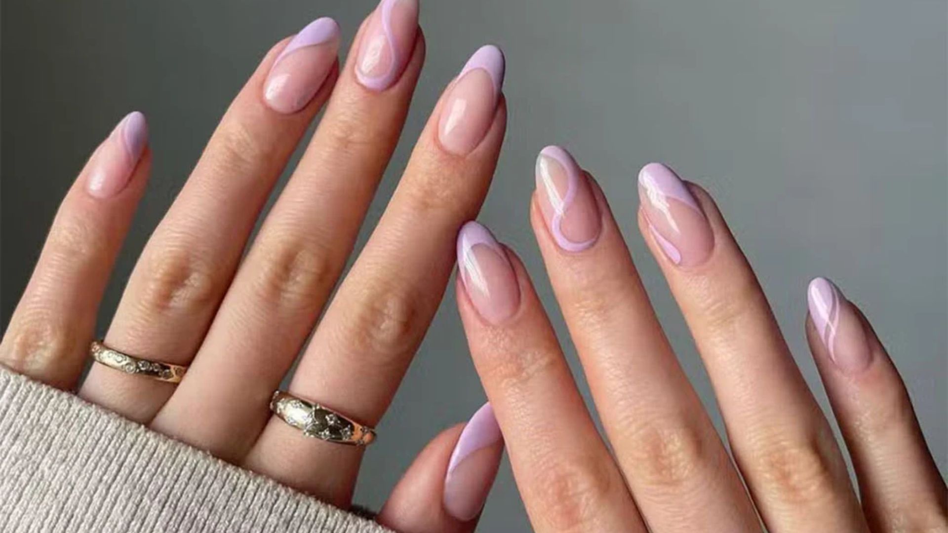 Close-up of manicured hands with almond-shaped nails, painted with a pastel lilac and white swirl design. Two fingers have gold rings. Cozy and elegant vibe.