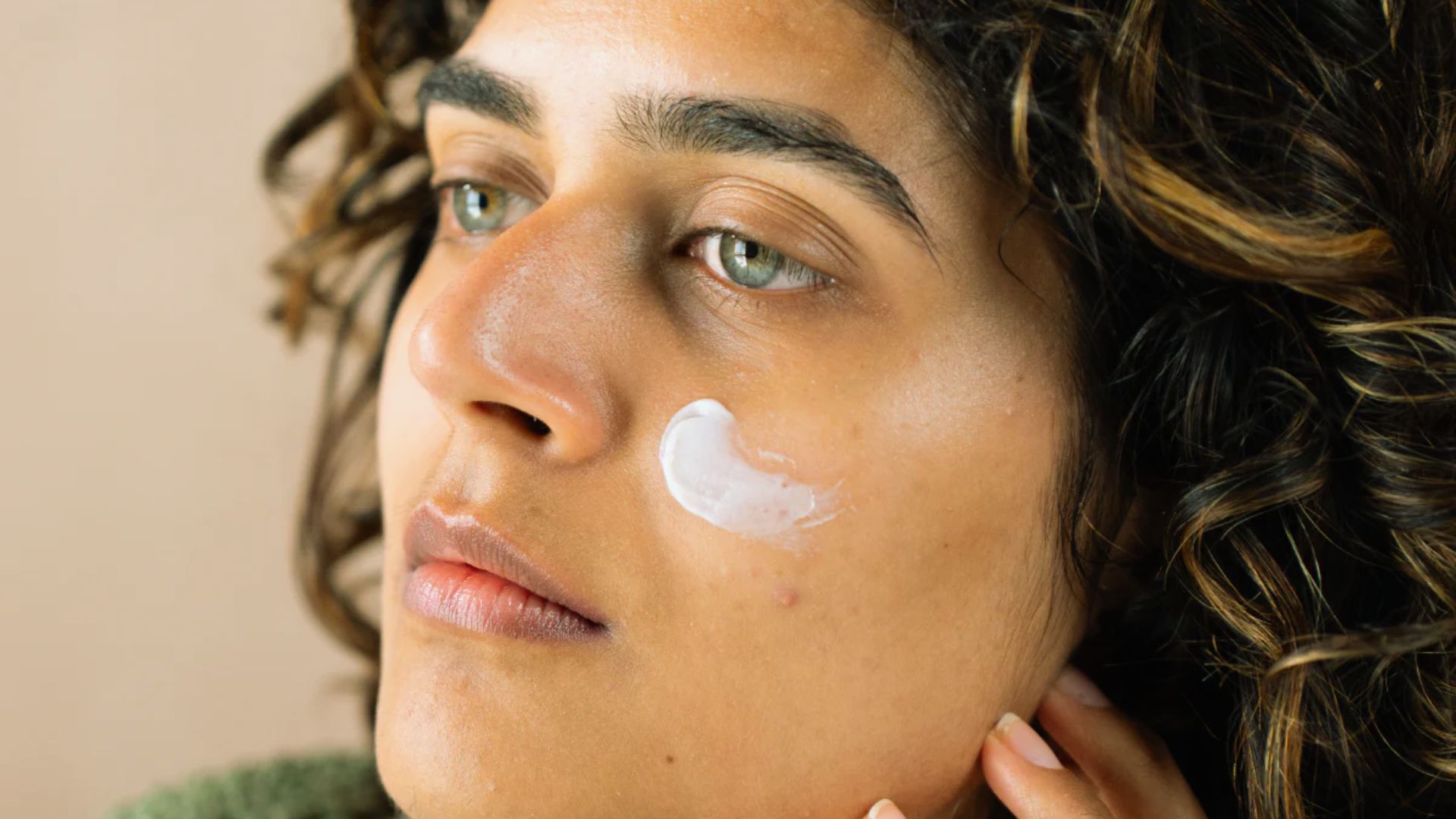 Person with curly hair applies moisturizer to their cheek, looking contemplative. The close-up highlights smooth skin and serene expression.