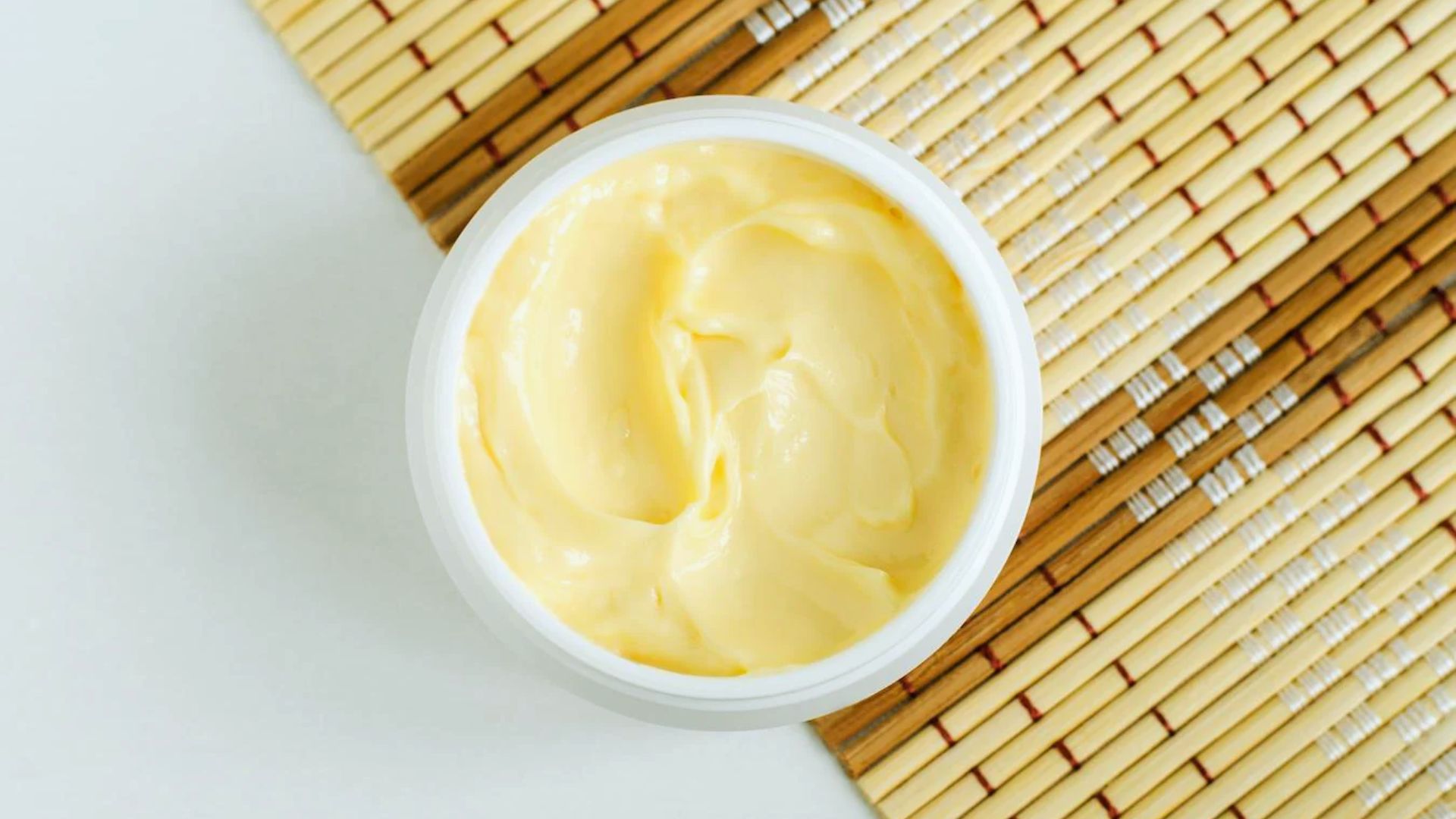 A round container of smooth, creamy yellow butter sits on a woven bamboo mat, creating a fresh and natural aesthetic.