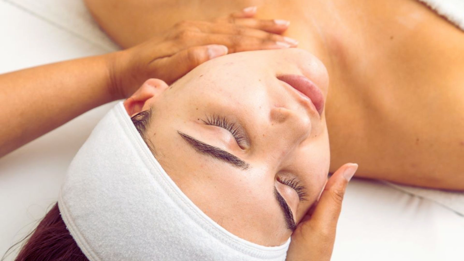 A person wearing a white headband receives a facial massage with eyes closed, relaxed. Hands gently cradle the face, conveying a serene spa experience.