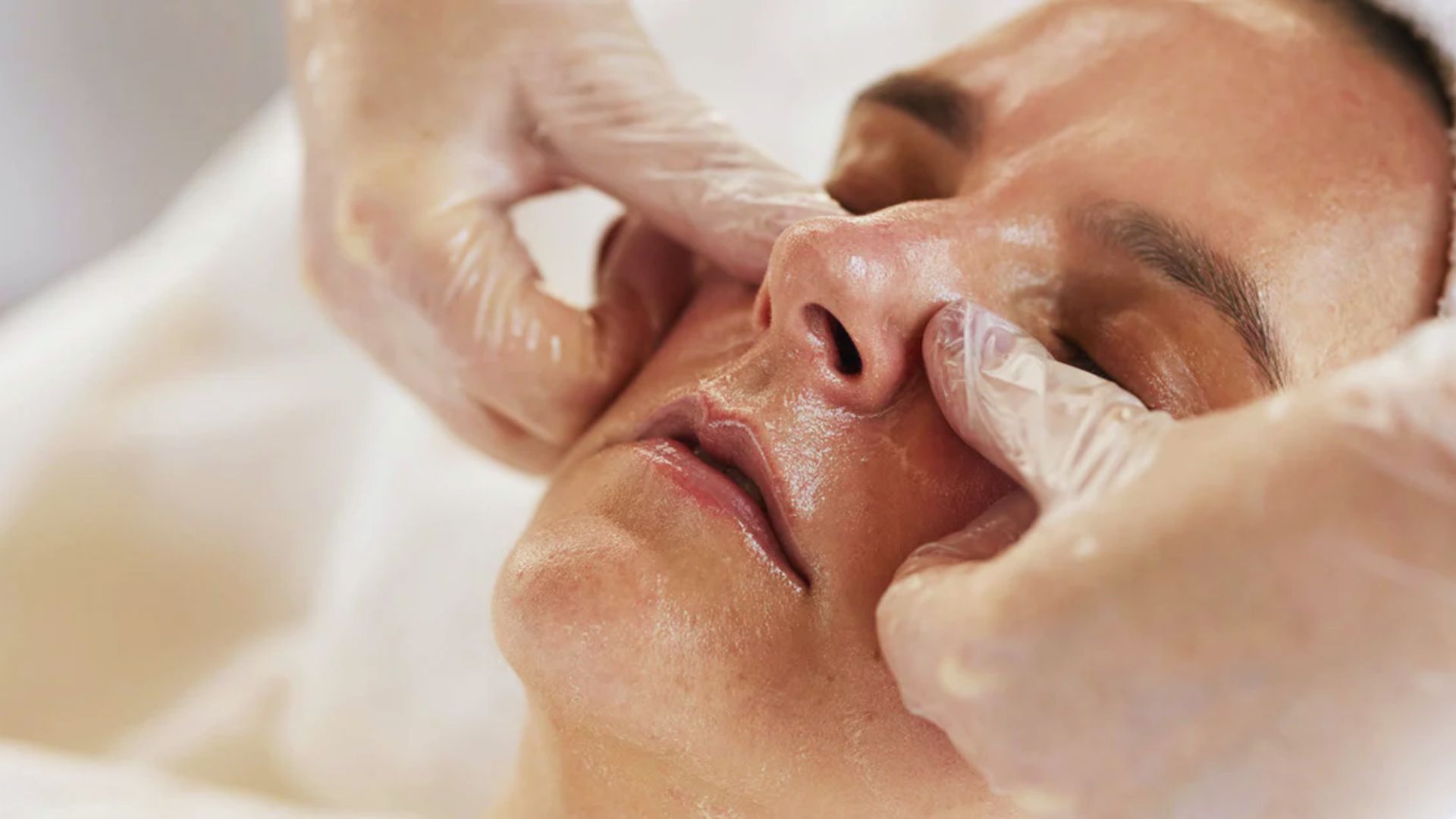 A close-up of a person's face receiving a facial massage. Hands in gloves gently press the cheeks, conveying relaxation and a sense of calm.