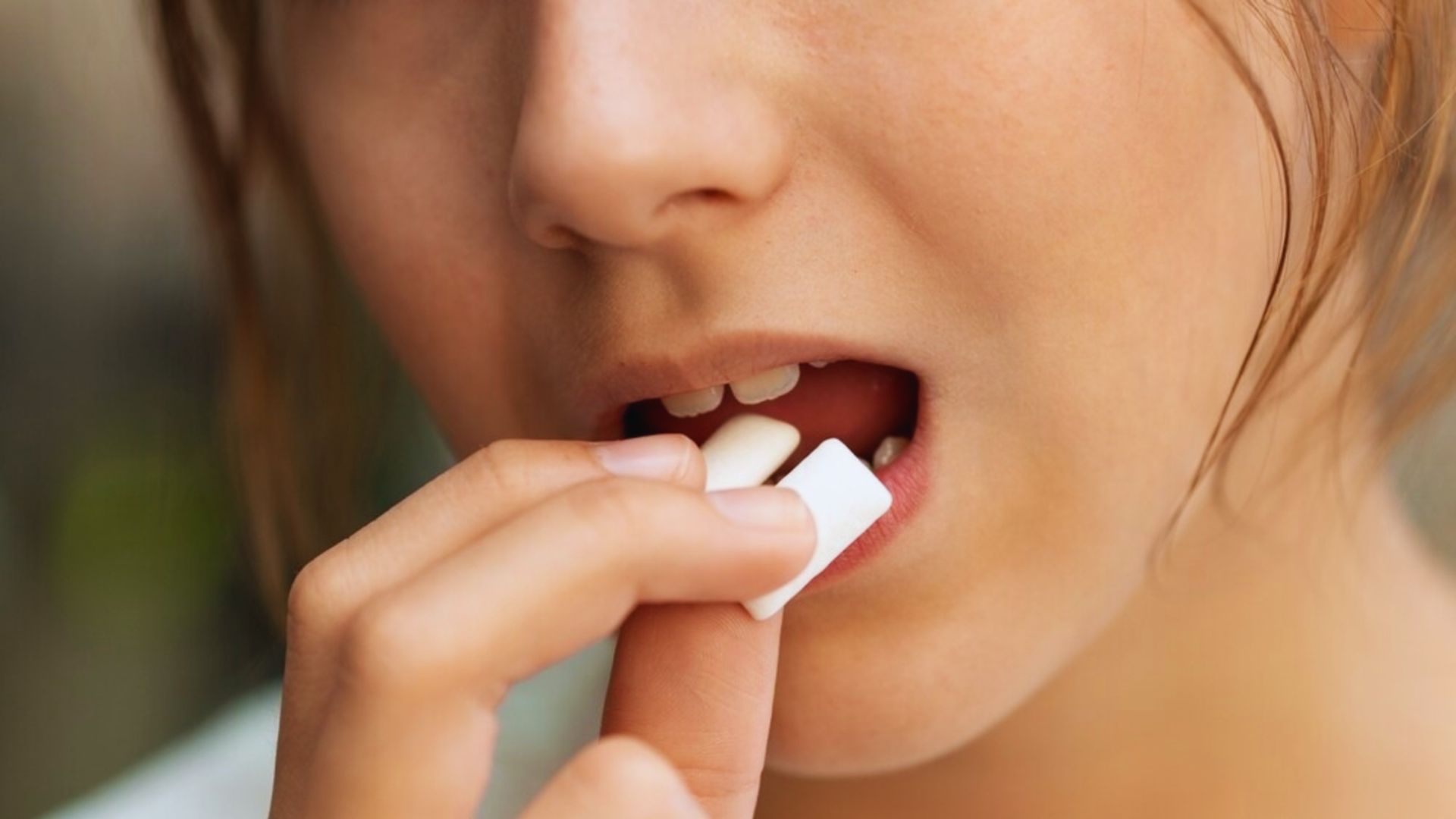 A close-up of a person placing chewing gum into their mouth. The focus is on the hand, mouth, and gum, conveying a casual and relaxed tone.