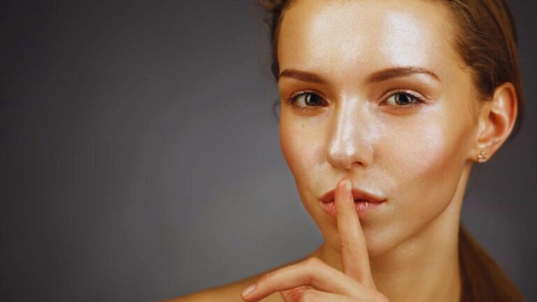 A woman with her finger to her lips in a "shh" gesture against a dark gray background, conveying secrecy or silence, with a serious expression.