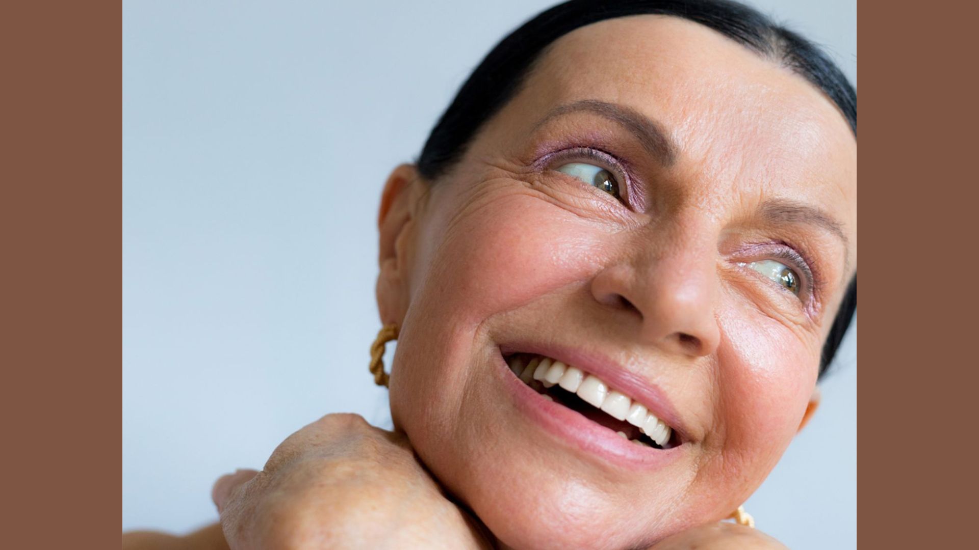 Elderly woman with short dark hair smiles joyfully, resting her chin on her hands. She exudes happiness against a soft blue background.