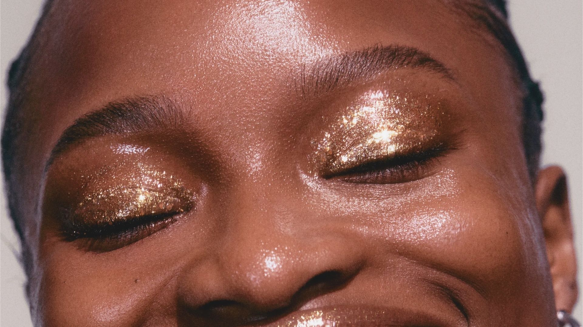 Close-up of a person with closed eyes, showcasing sparkling gold eyeshadow. The expression conveys joy and confidence, emphasizing a radiant, glamorous look.