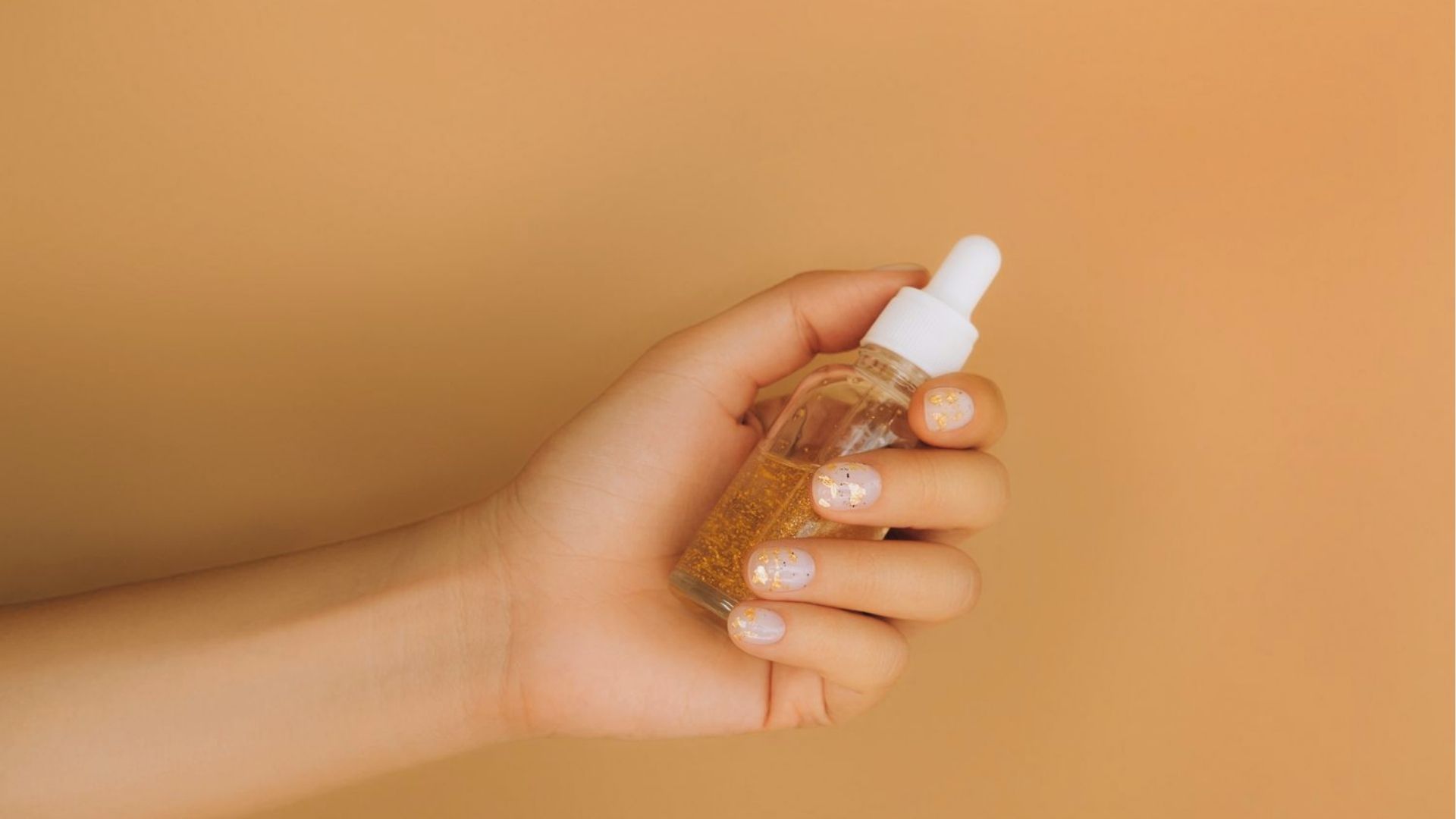 A hand with painted nails holds a dropper bottle filled with a gold liquid against a warm beige background, conveying a sense of elegance and beauty.