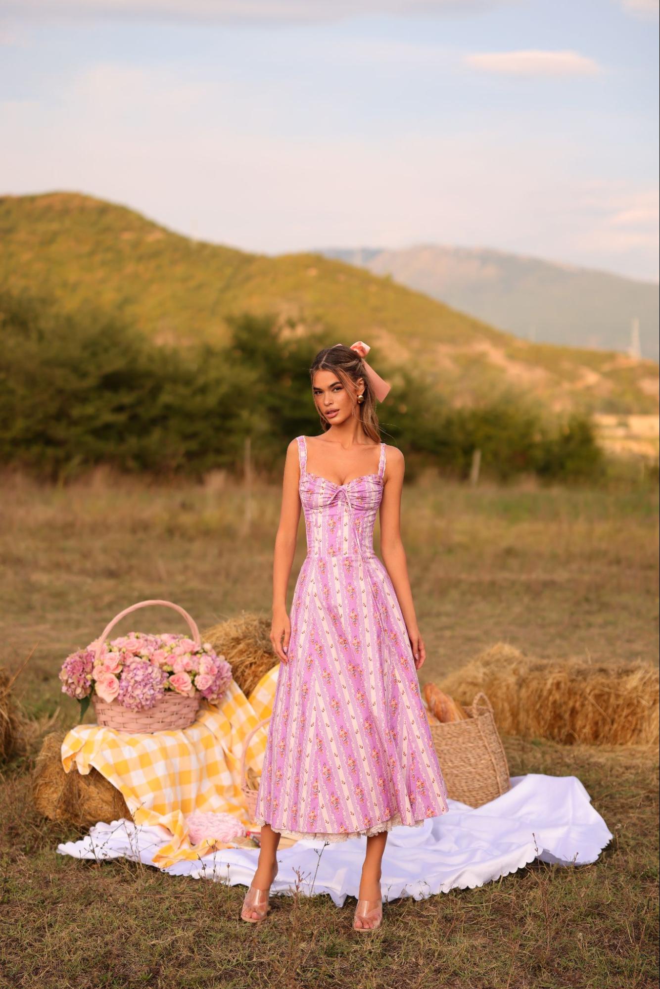A woman in a pink dress stands on a picnic blanket, surrounded by a serene outdoor setting.