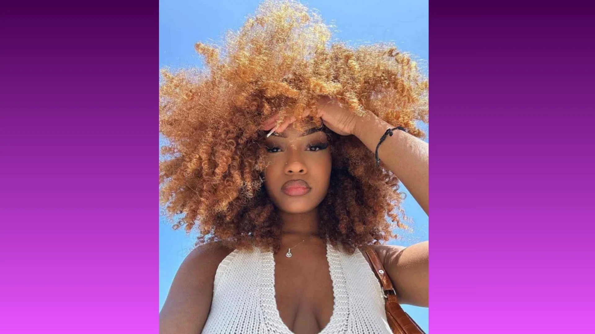 Woman with voluminous curly hair holds hand to forehead, gazing up. She wears a white knit top against a clear blue sky, exuding confidence.