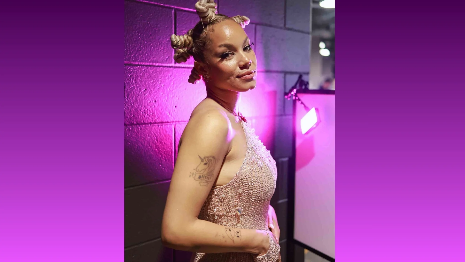 A person with braided hair in a sparkling pink dress stands confidently against a purple-lit wall, exuding elegance and confidence.