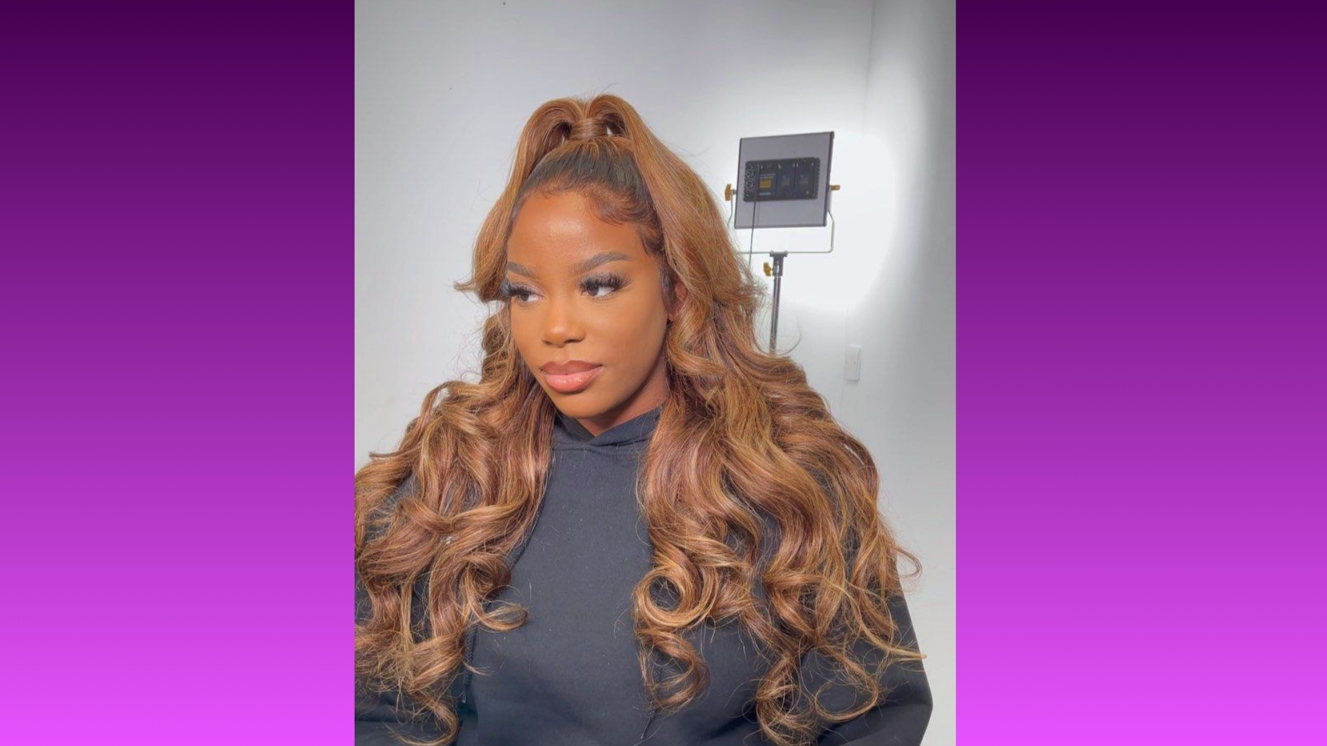 Young woman with long, curly, light brown hair styled in a high ponytail, wearing a black hoodie. She's lit warmly and framed by a gradient purple background.