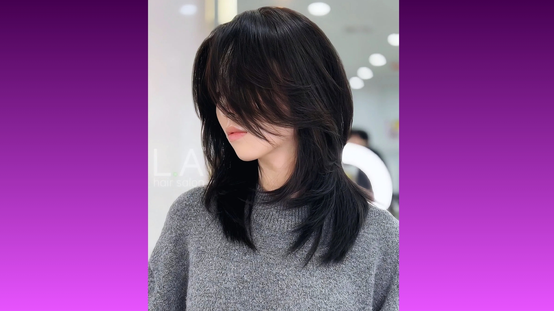 Young woman with long, layered black hair and curtain bangs, wearing a gray sweater. She's indoors against a blurred background, exuding a calm vibe.