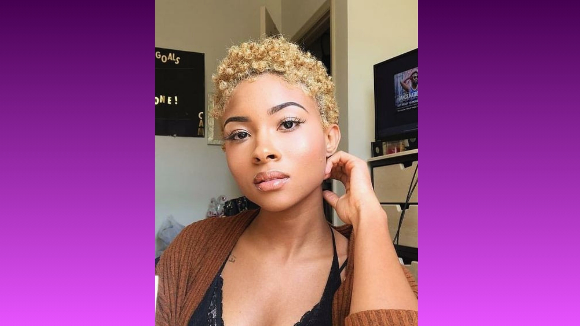 A person with short, curly blonde hair and natural makeup poses indoors with a relaxed expression. A TV and a "Goals" board are in the background.