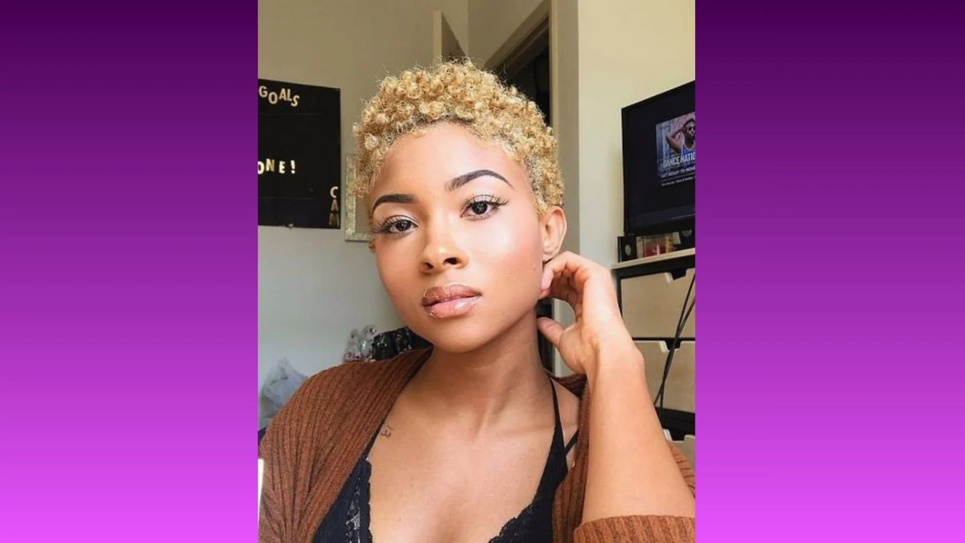 A person with short, curly blonde hair and natural makeup poses indoors with a relaxed expression. A TV and a "Goals" board are in the background.
