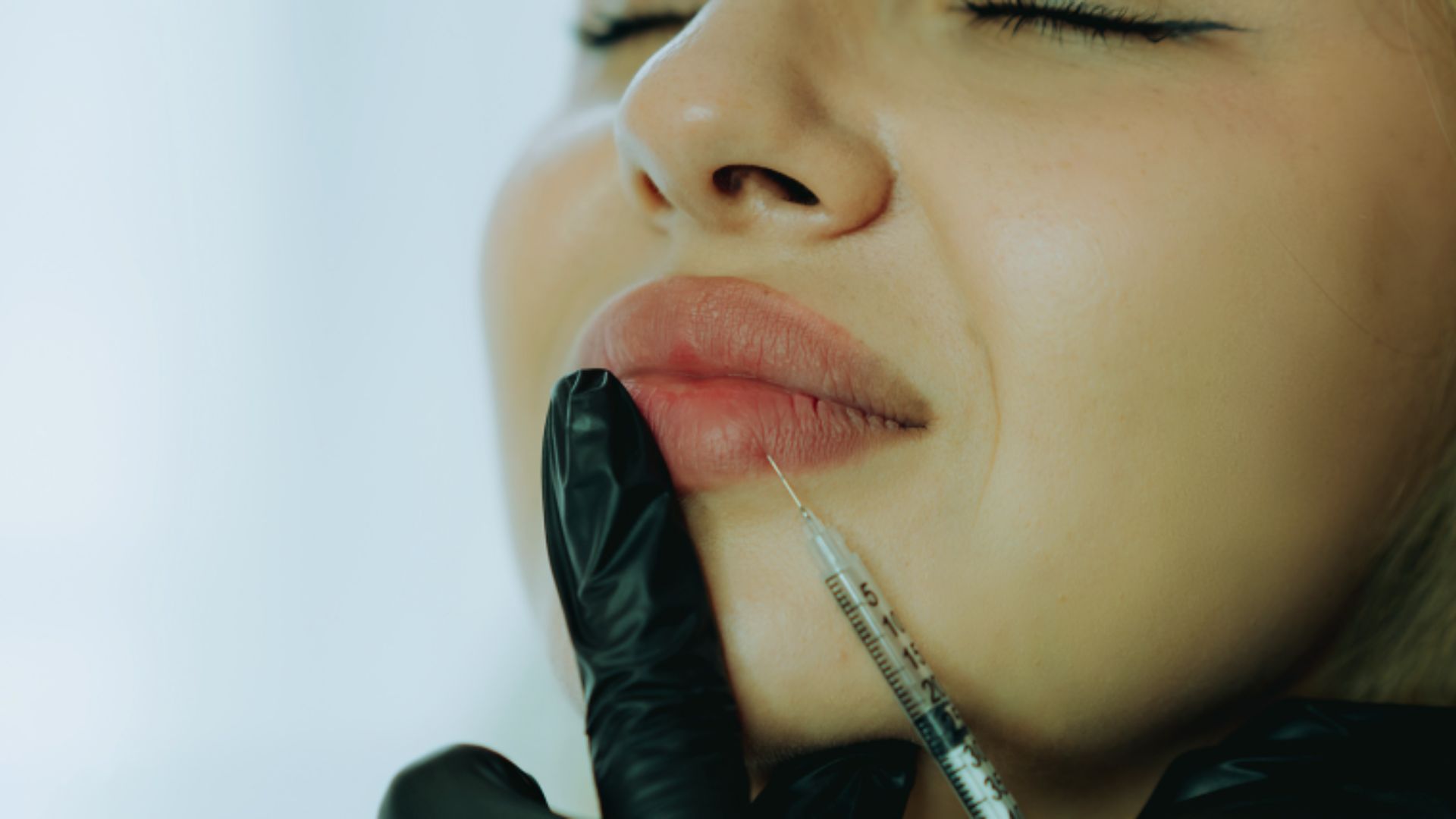A woman with closed eyes receives a lip injection from a gloved hand, conveying a sense of calm. Her lips are slightly pursed.