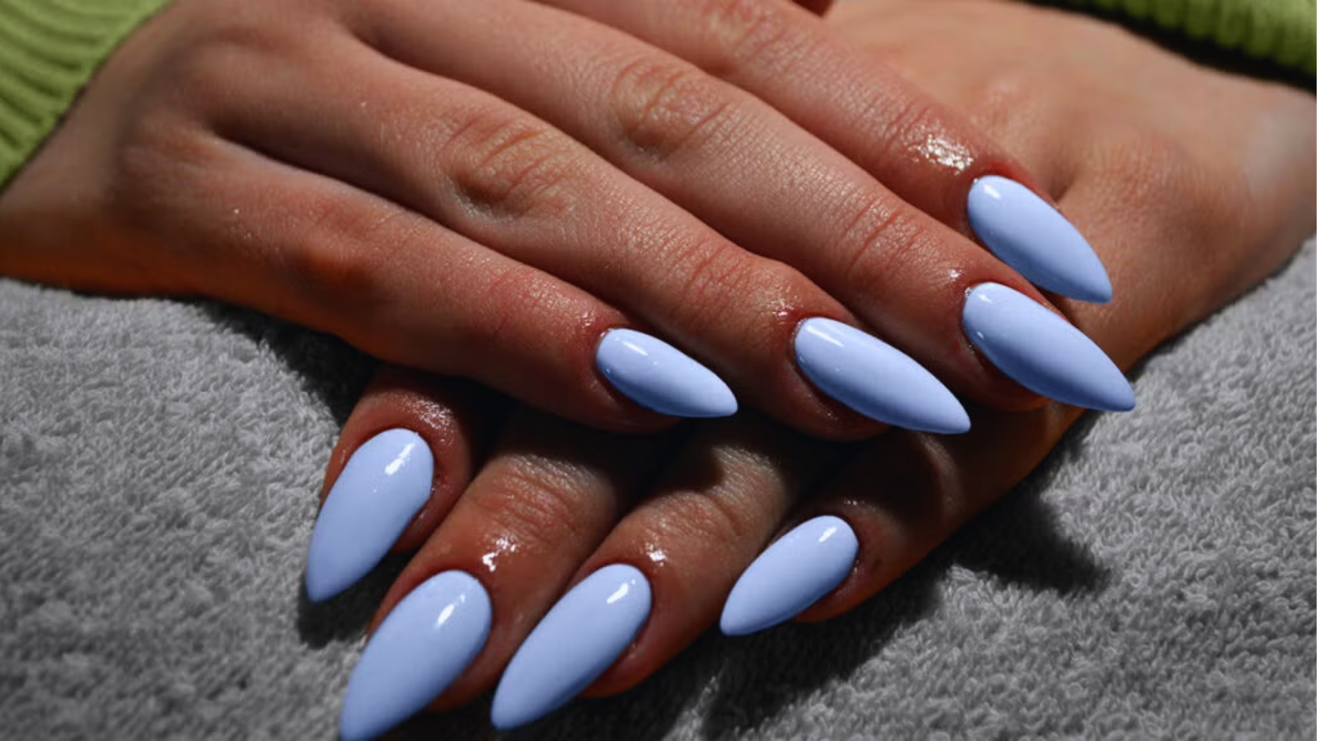 Hands with long, almond-shaped light blue nails resting on a gray towel. The setting is calm and elegant, with a soft, cozy ambiance.