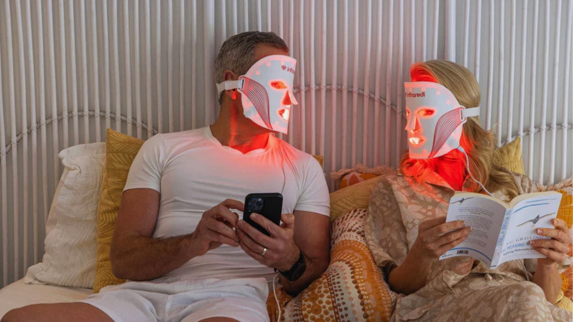 A couple wearing LED light therapy masks sits together on a bed. One holds a smartphone, the other a book. The setting is cozy and relaxed.