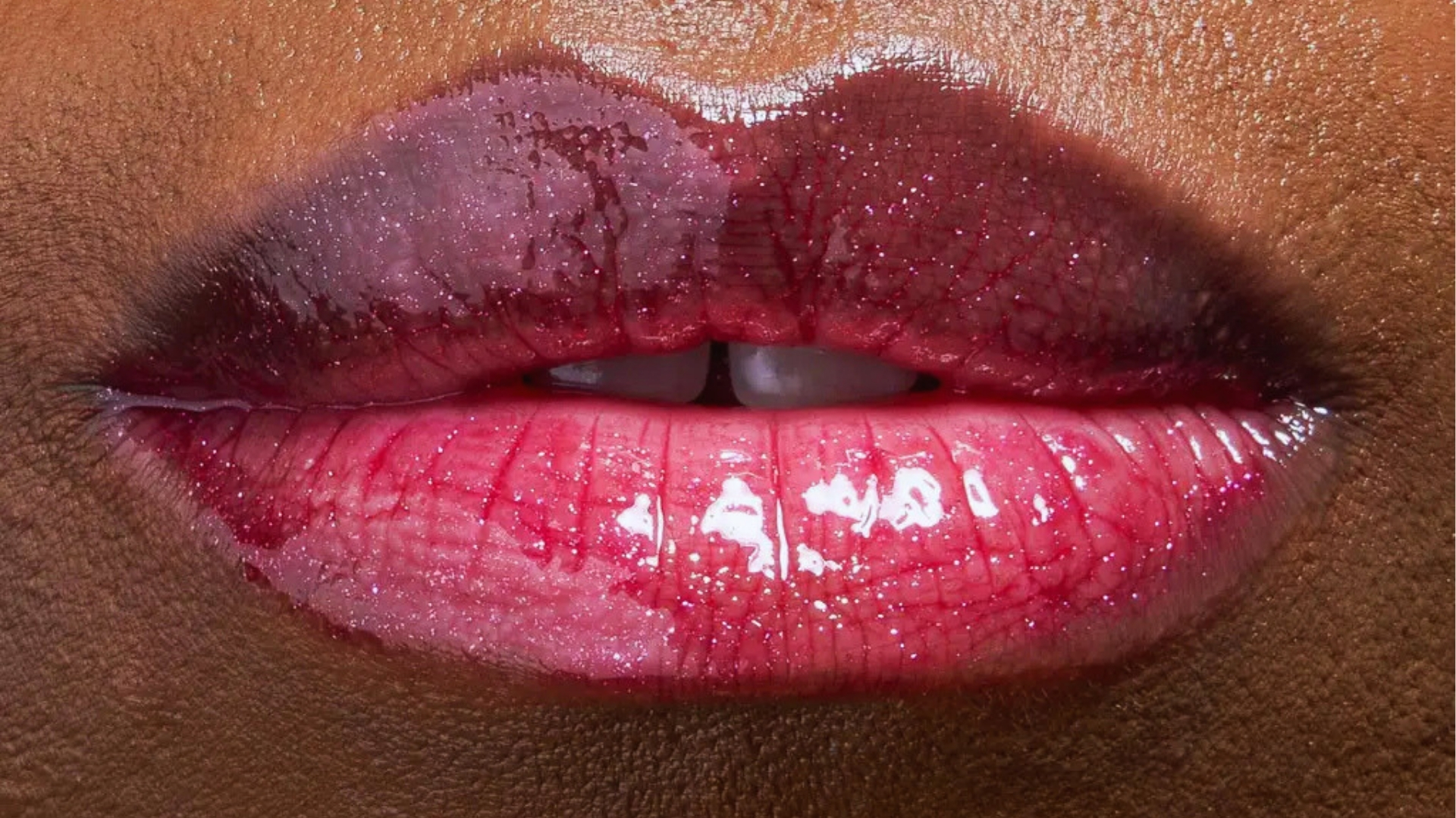 Close-up of glossy lips with a two-tone effect; upper lip is dark purple, lower lip is bright pink. The texture is shiny and reflective.