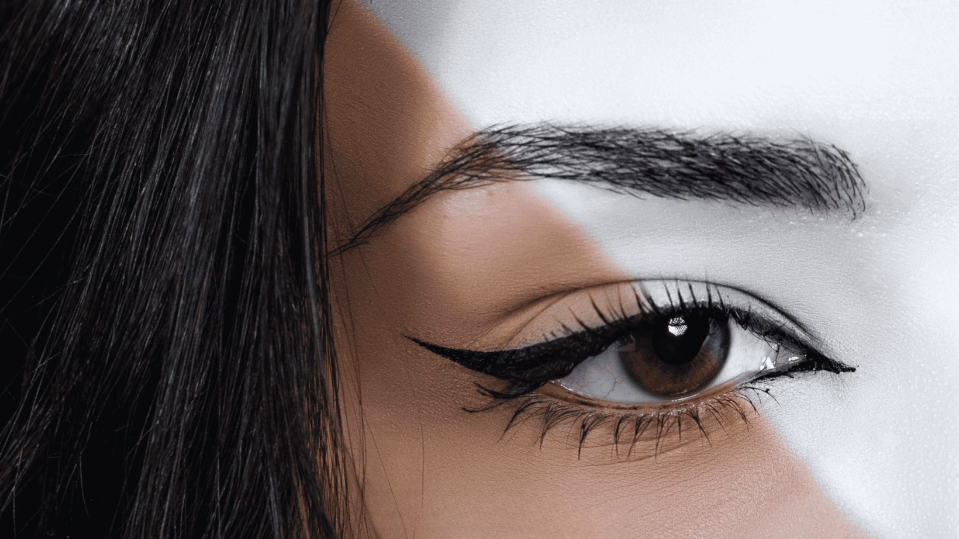Close-up of a woman's eye with bold eyeliner, divided diagonally by color; one side in natural tones, the other in grayscale, evoking contrast.