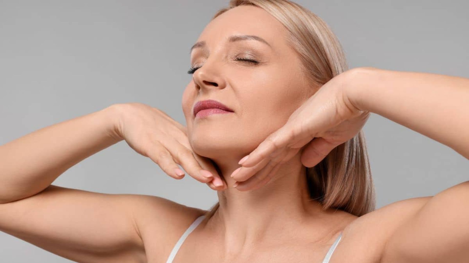 A relaxed woman with closed eyes tilts her head slightly up, with hands gently under her chin against a gray background, conveying tranquility.