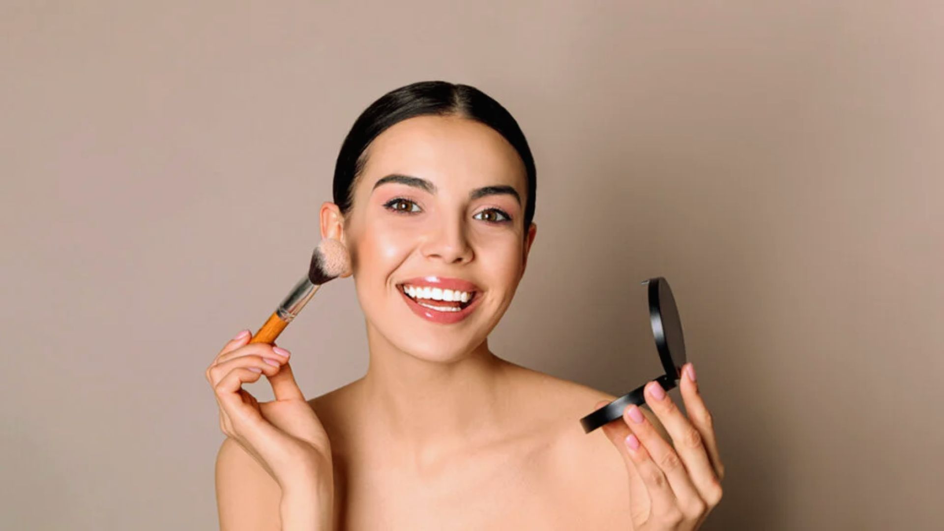 Smiling woman applying makeup with a brush, holding a compact. Background is plain beige. She exudes confidence and joy, with glowing skin.