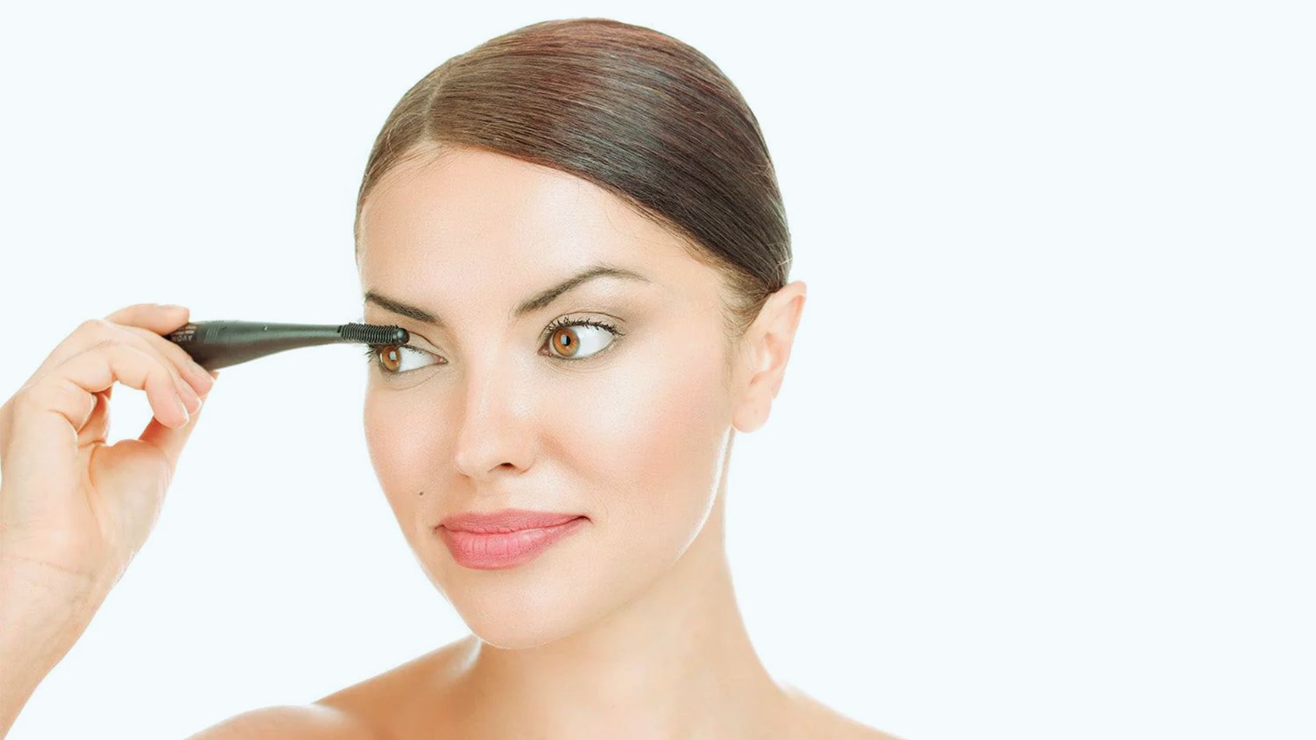 A woman with a smooth complexion and hair pulled back applies eyeliner with concentration. The mood is calm and focused against a plain background.