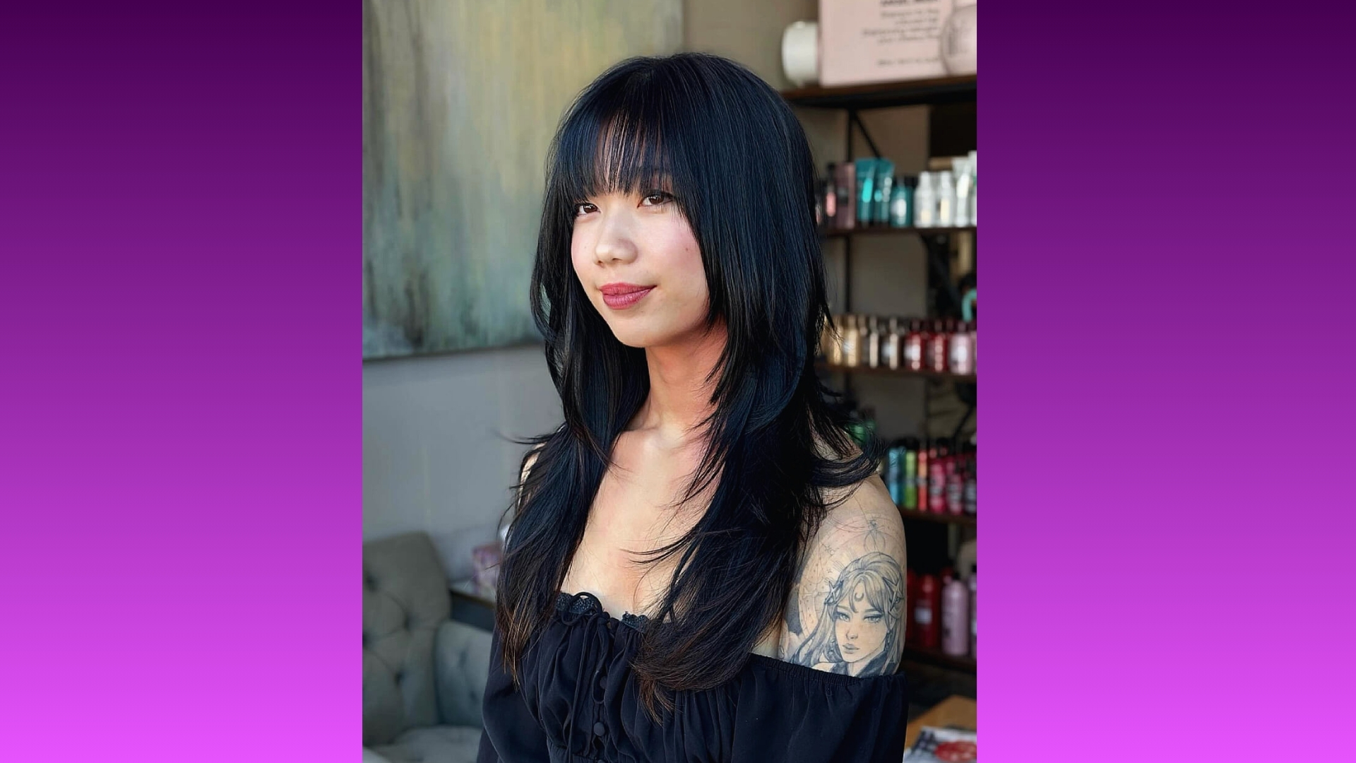 A woman with long, layered black hair and bangs poses indoors. She wears an off-shoulder black top, revealing a detailed tattoo on her arm. Shelves behind her display various products, contributing to a relaxed, stylish atmosphere.