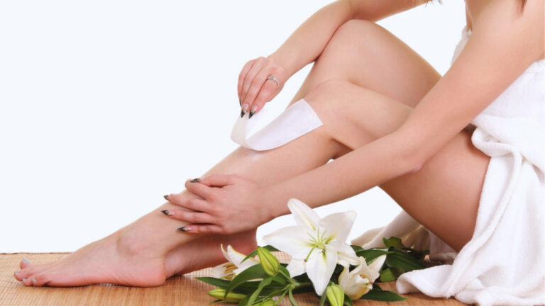 A woman wrapped in a white towel gently waxes her leg, surrounded by fresh white lilies. The setting is calm and spa-like, suggesting relaxation.
