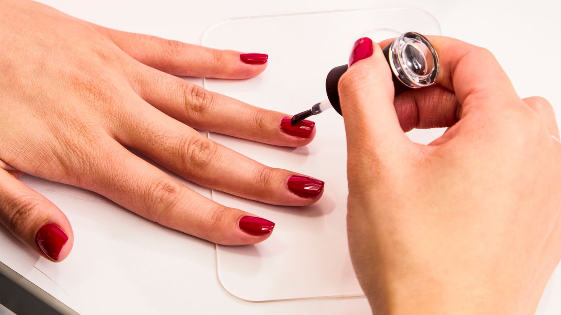 A person applies glossy red nail polish to neatly manicured nails on another's hand, conveying a sense of care and elegance.