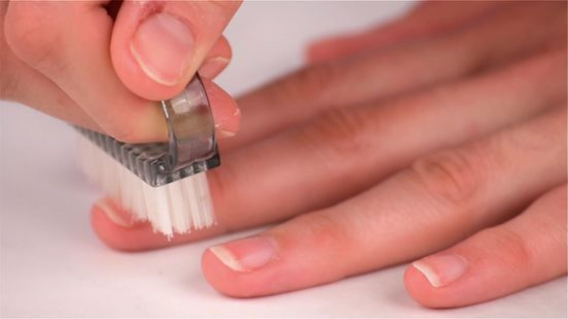A hand uses a small brush to clean fingernails on the other hand. The background is a soft white, conveying a hygienic, grooming-focused scene.