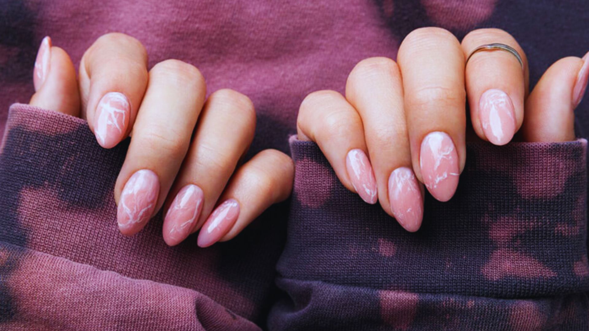 Close-up of hands with pink marble-patterned nails, resting on a maroon tie-dye fabric. The nails are almond-shaped, conveying a stylish and trendy vibe.