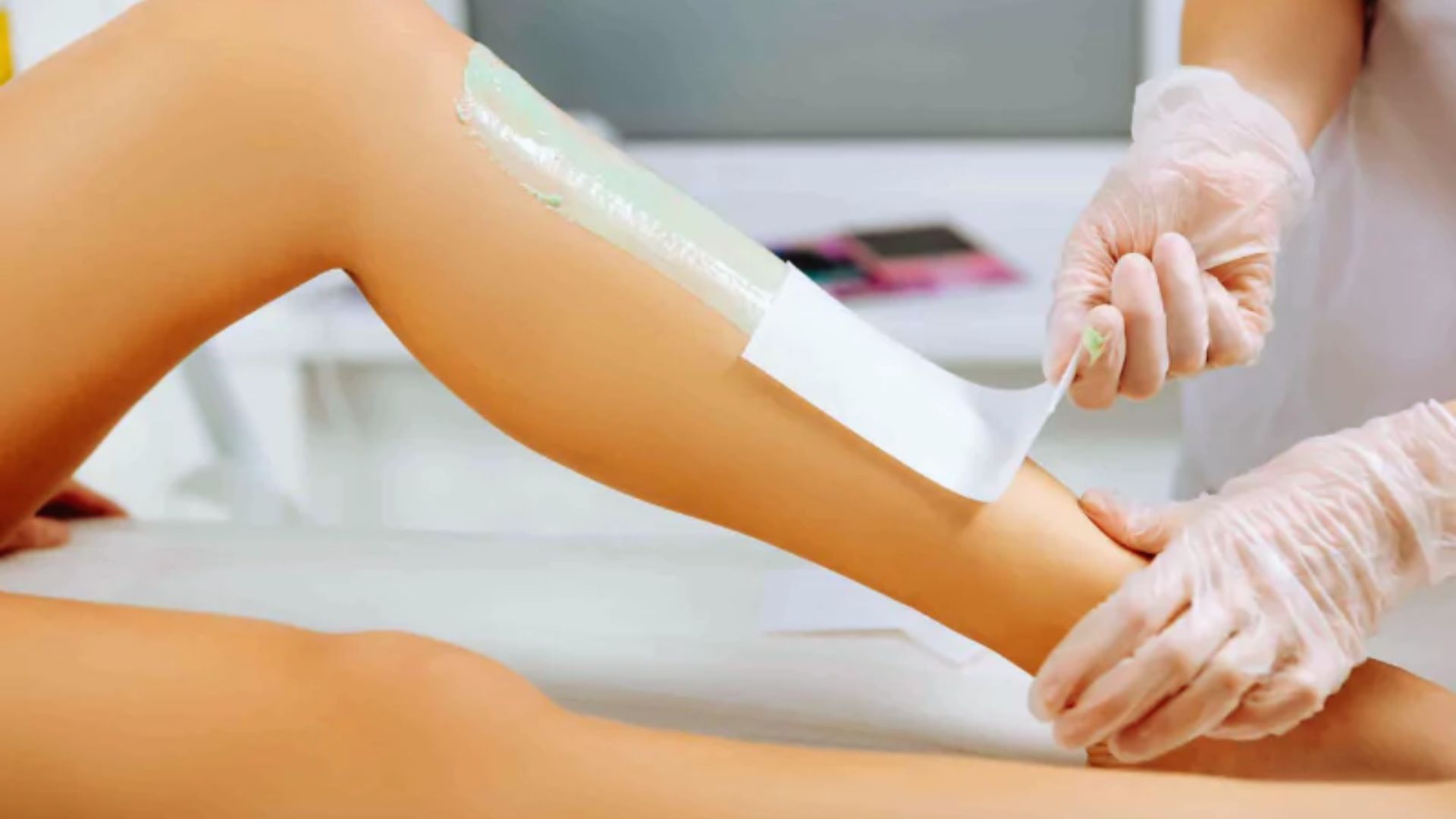 A person receiving a leg waxing treatment. A strip is being applied over green wax by gloved hands, suggesting a calm, professional setting.