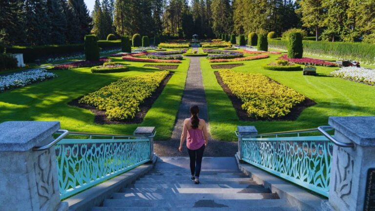 A woman in a pink top descends ornate stairs toward a vibrant, symmetrical garden with colorful flowerbeds and manicured lawns, surrounded by tall trees.