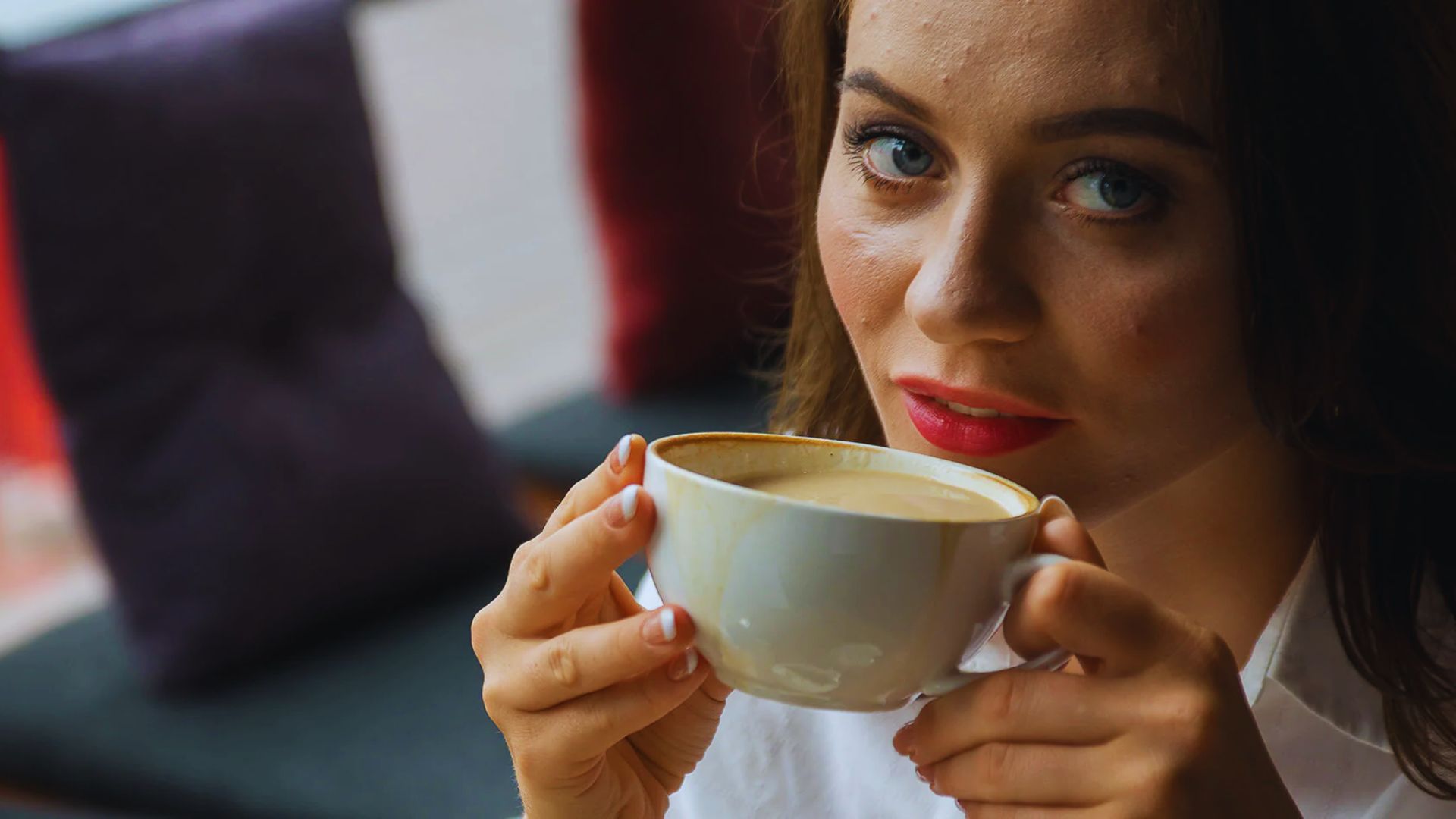 Woman with red lipstick and blue eyes holds a white coffee cup near her face, gazing intently. Cozy setting with soft pillows in the blurred background.