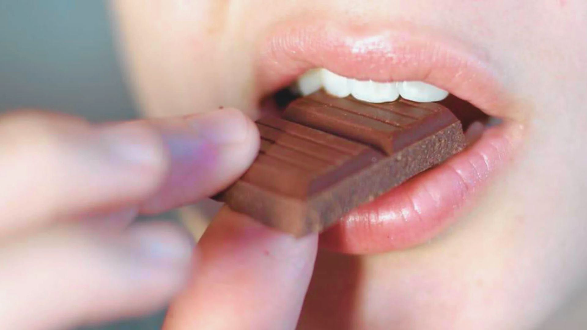 Close-up of a person about to bite a piece of chocolate, with lips slightly parted and fingers holding the chocolate. The scene conveys anticipation and indulgence.