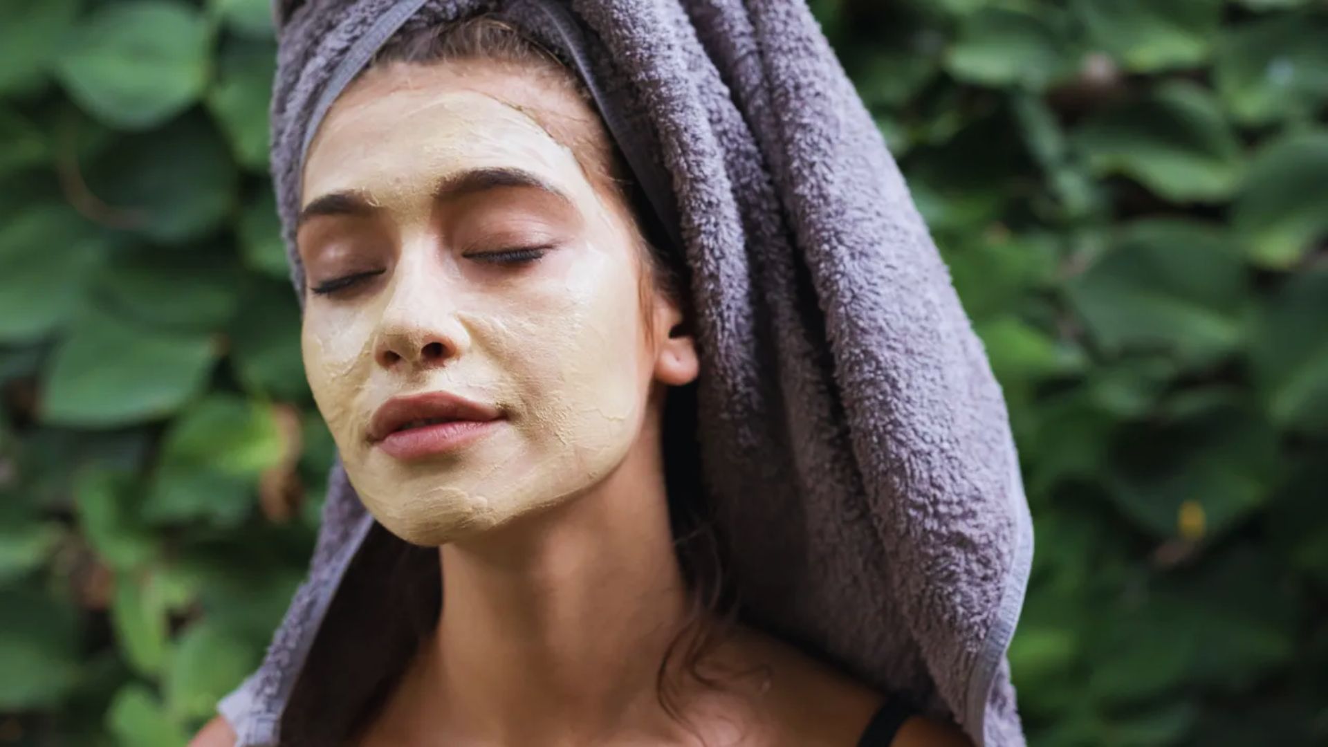 Woman with a towel wrapped around her head enjoys a facial mask, eyes closed, against a lush green leafy background, conveying relaxation and tranquility.