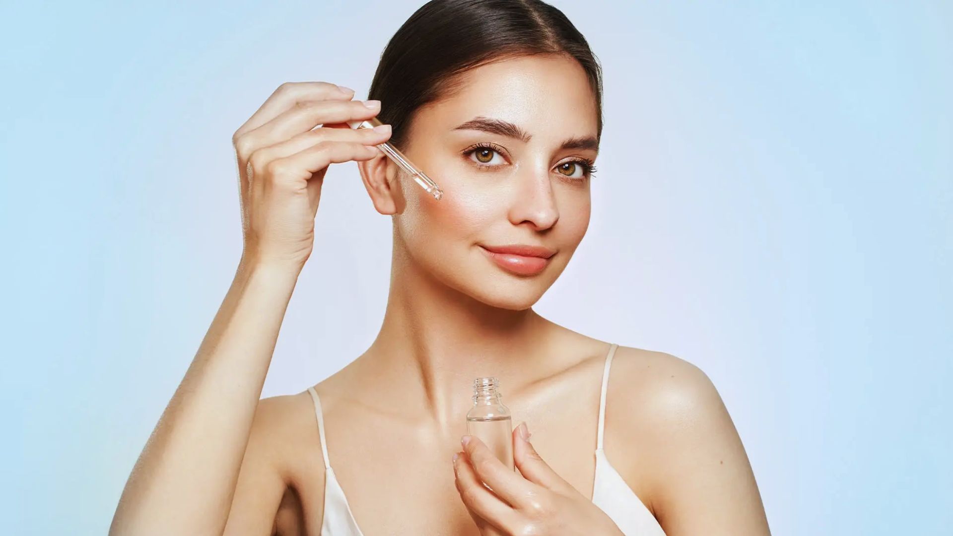 A woman with clear skin smiles while applying facial serum using a dropper. She wears a white tank top against a light blue background, conveying a fresh, healthy vibe.