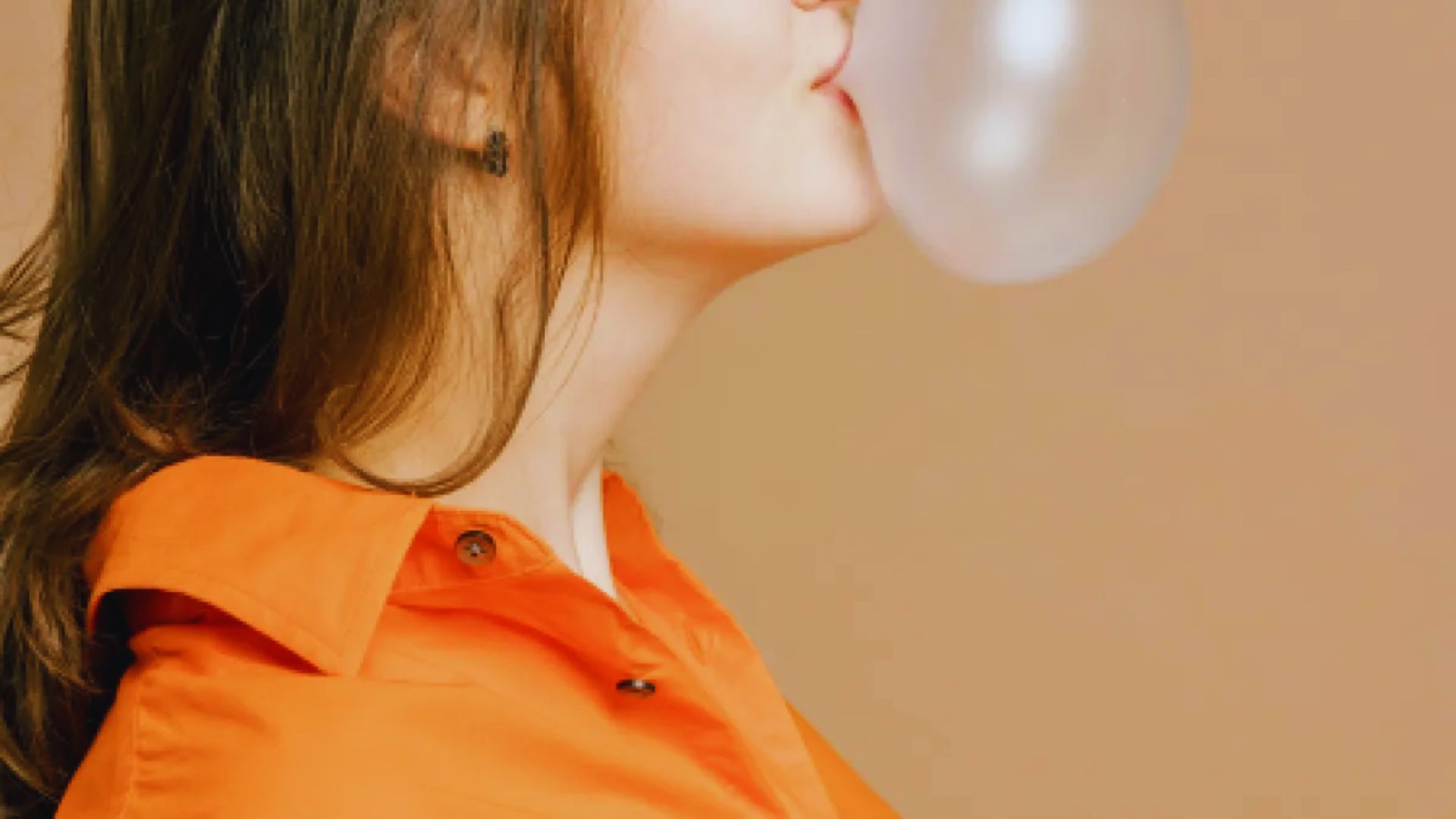 A person in an orange shirt blows a large bubble with gum against a beige background. The mood is playful and lighthearted.