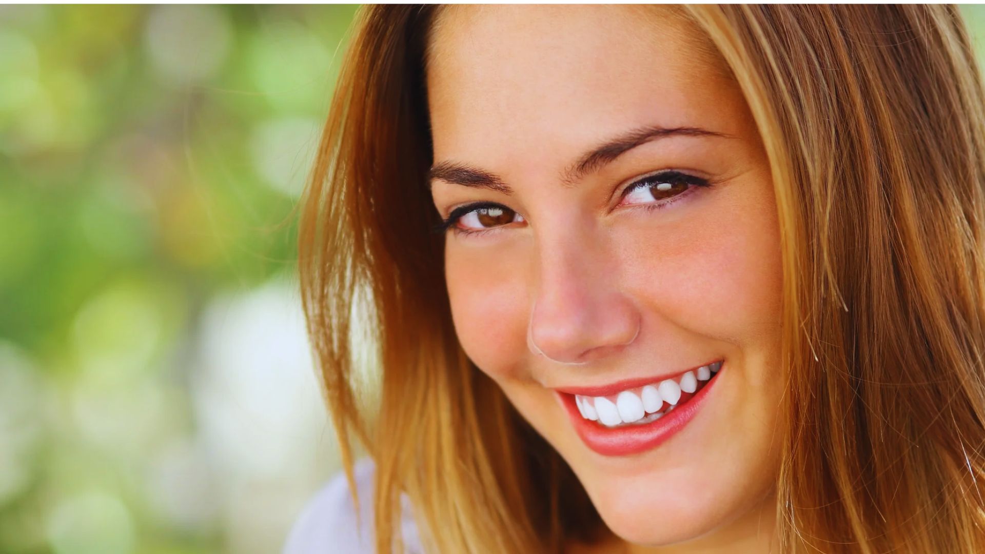 Smiling young woman with long brown hair, showing straight white teeth. Background is softly blurred with vibrant green tones, conveying a cheerful mood.