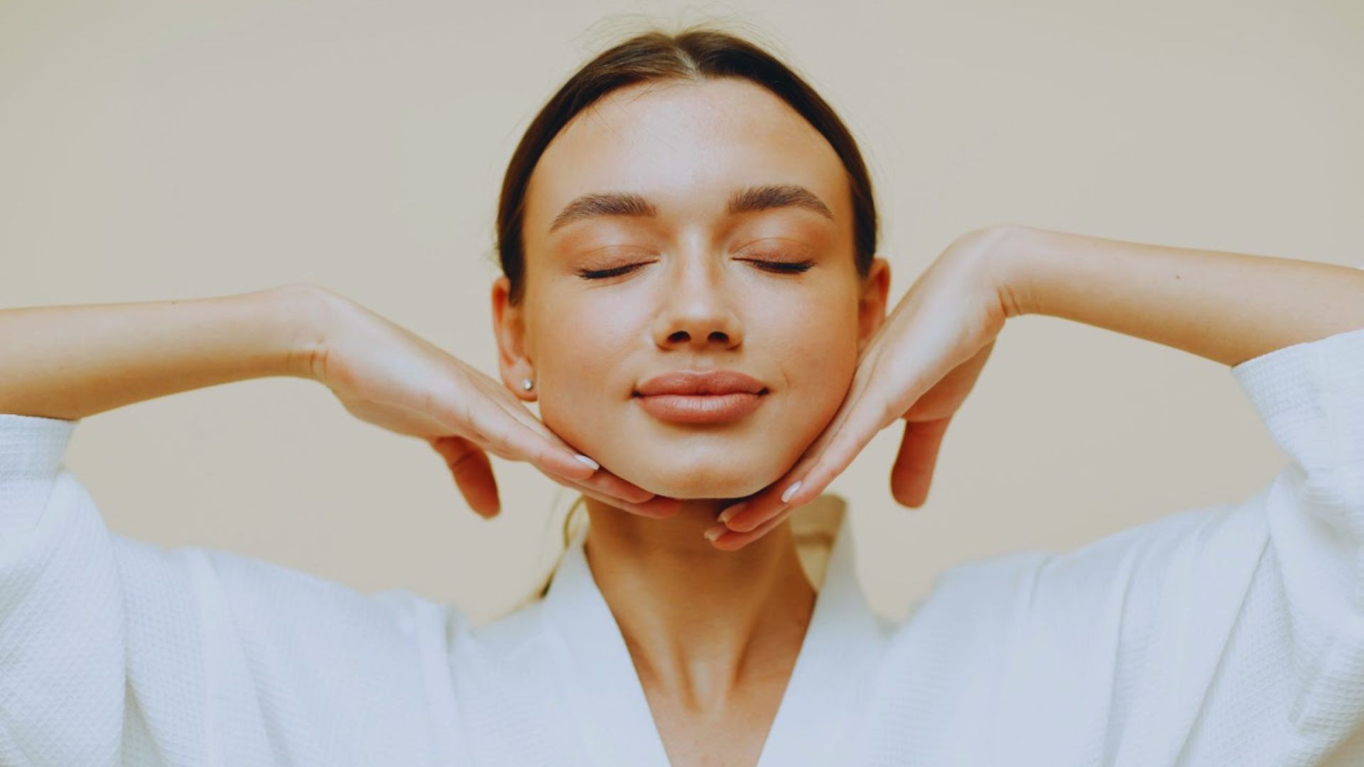 Young woman with eyes closed, gently lifting her chin with both hands in a relaxed pose. She wears a white robe against a neutral background, conveying calmness.