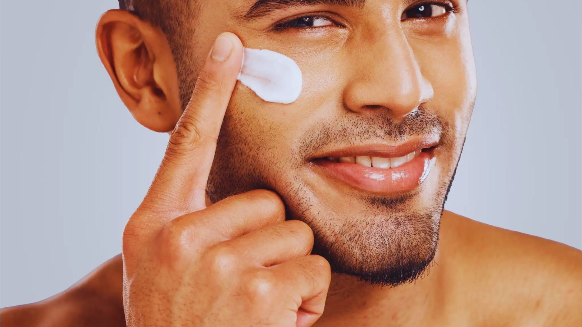 Man smiles while applying face cream with his finger on his cheek. The background is light blue, conveying a fresh and clean feeling.