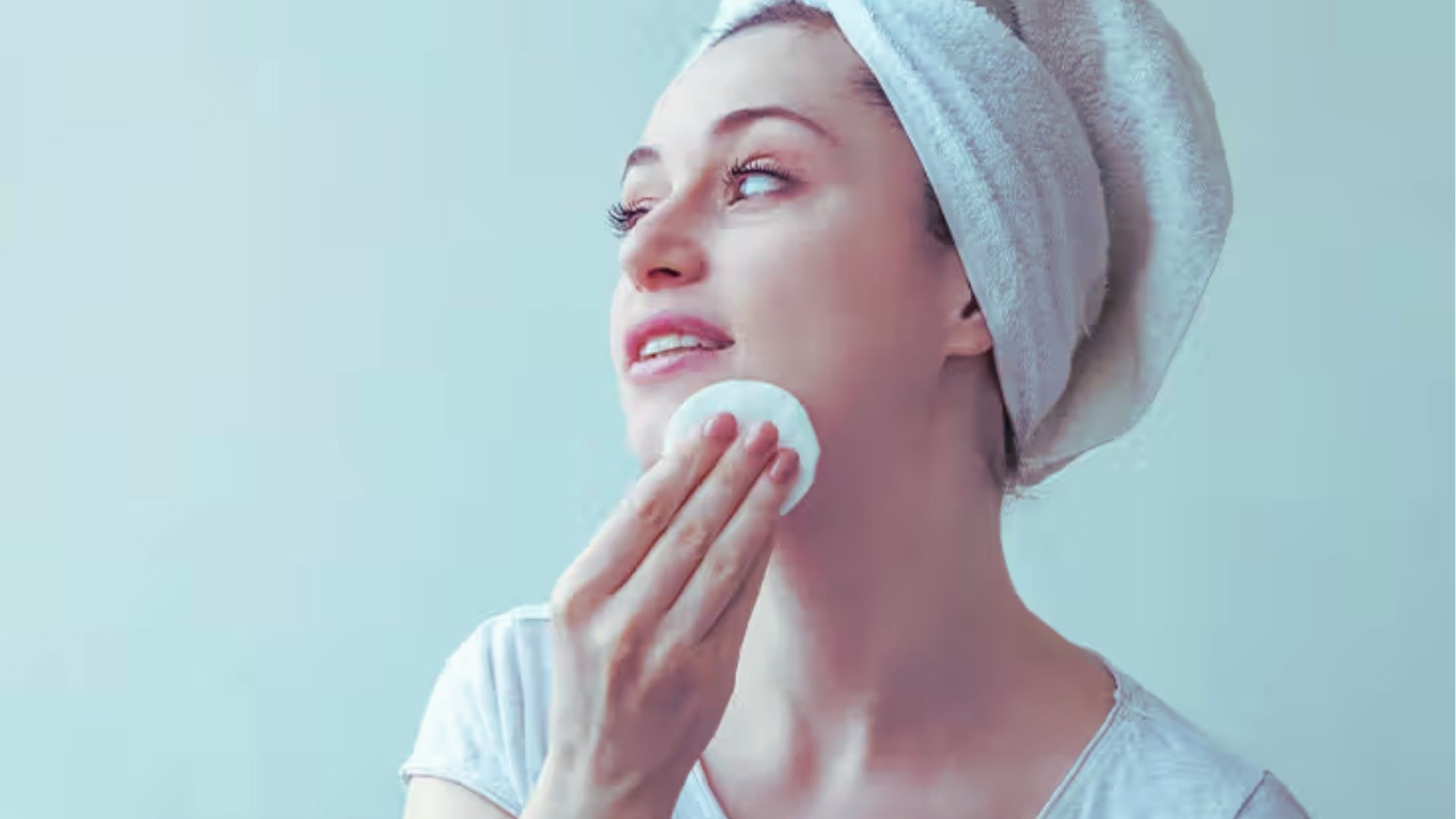 A woman with a towel wrapped around her head gently applies a cotton pad to her cheek, conveying a calm and refreshing skincare routine.