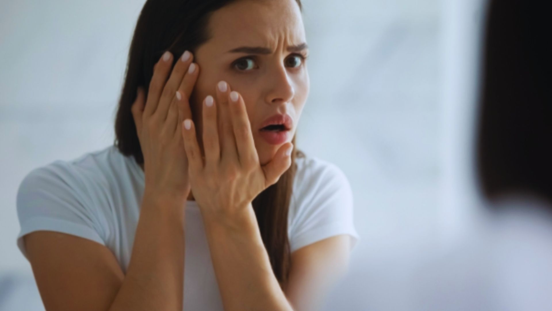 A woman in a white shirt looks into a mirror with surprise and concern, her hands on her cheeks. The background is softly blurred, emphasizing her expression.