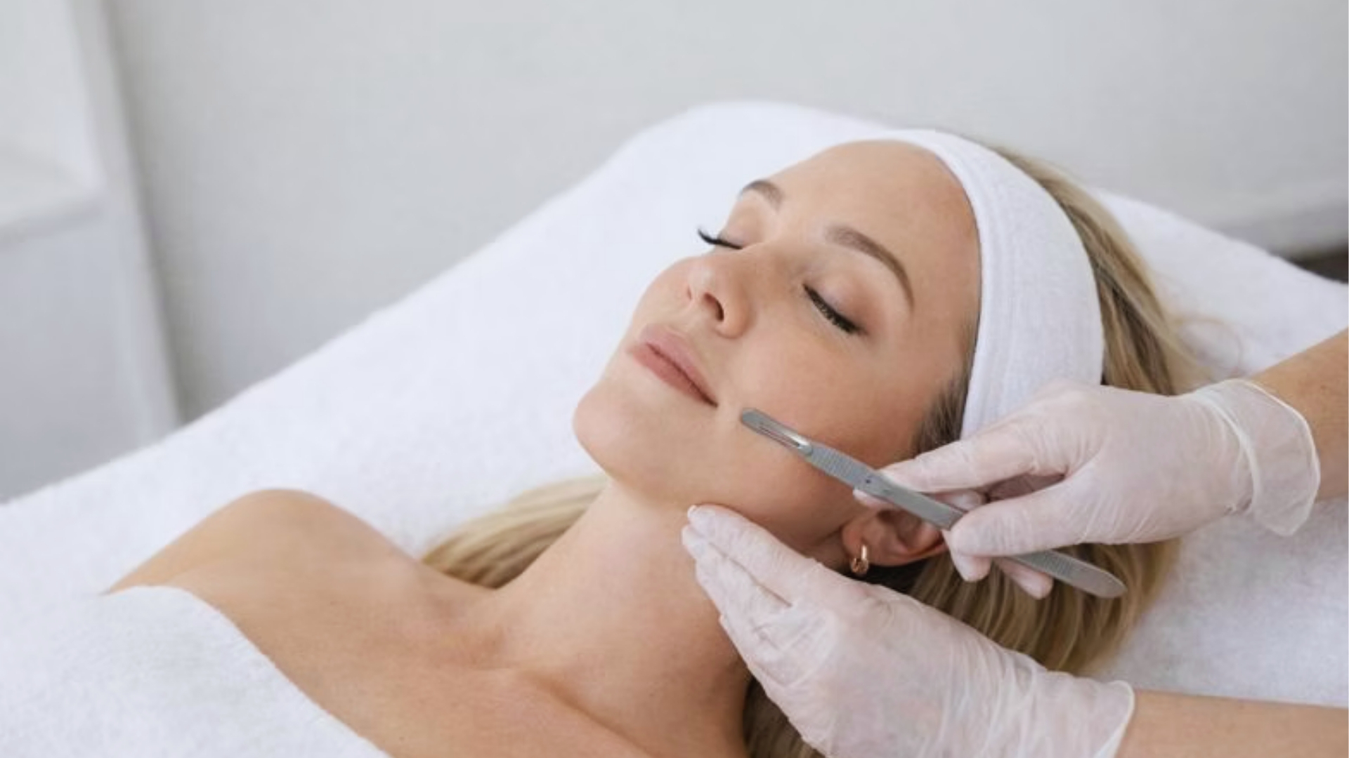 A woman lies relaxed on a spa table with eyes closed, receiving a dermaplaning treatment. Aesthetician's gloved hands hold a scalpel on her cheek.