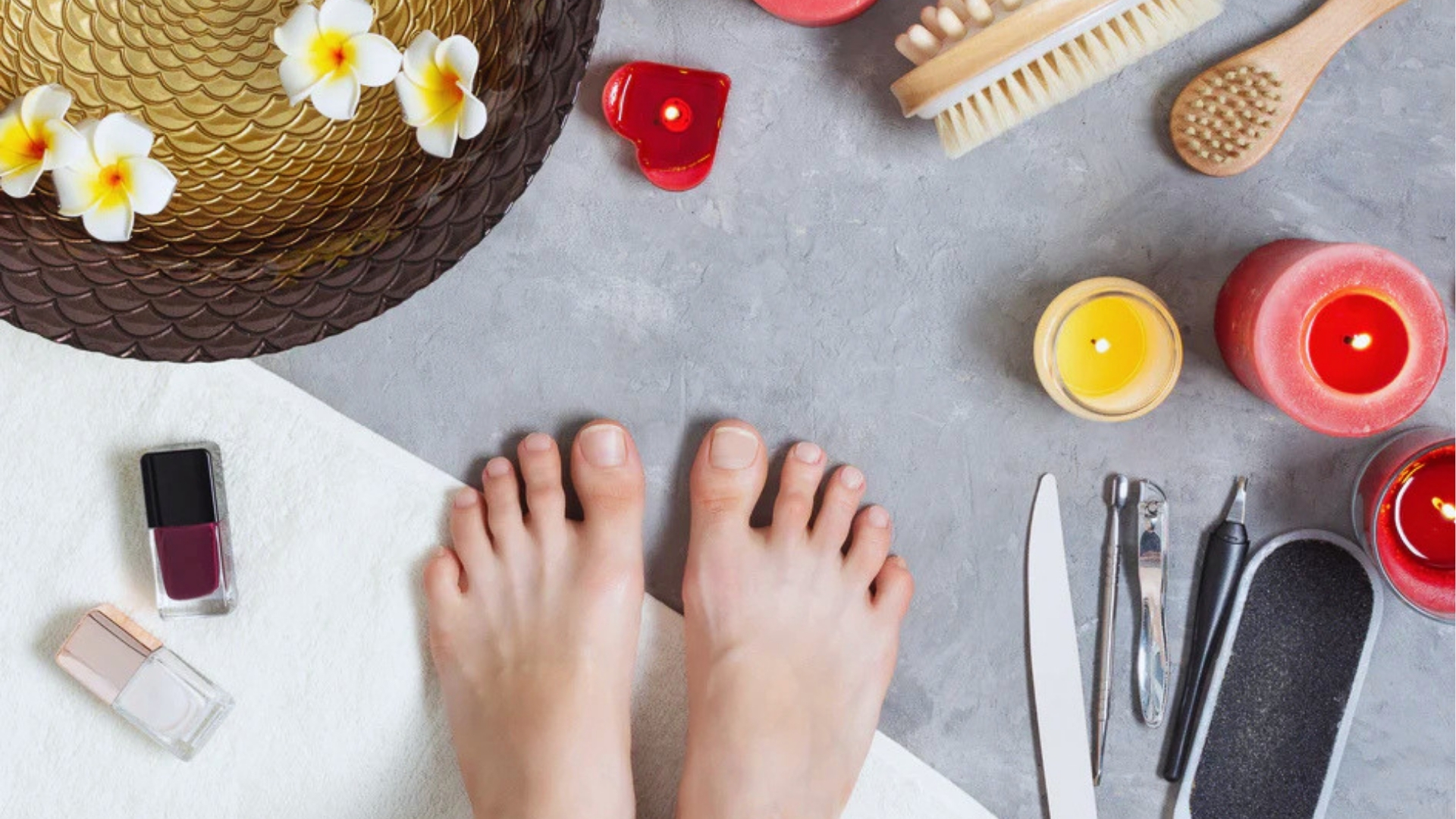 Bare feet on a towel, surrounded by nail polish, candles, spa tools, and frangipani flowers, set on a gray surface, conveying a relaxing spa atmosphere.