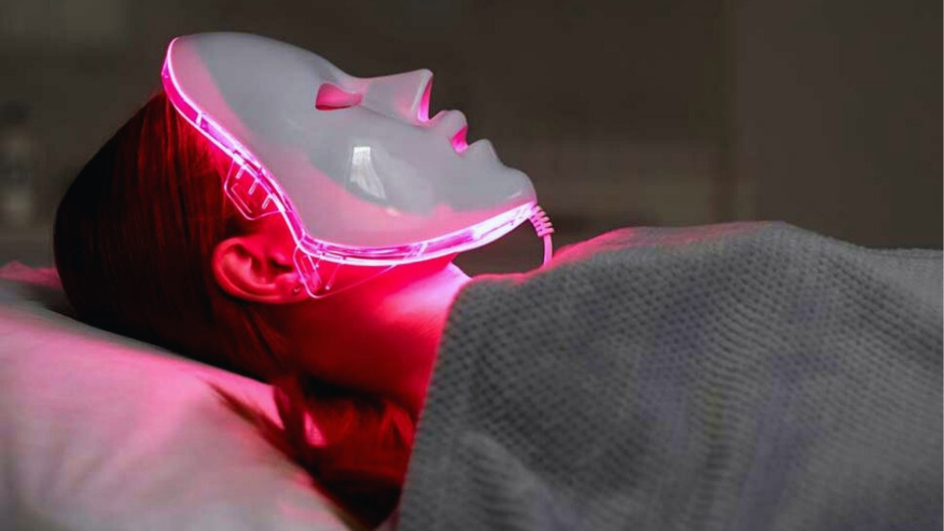 Person lying down wearing a glowing LED facial mask emitting pink light, creating a futuristic and calming atmosphere. The background is dim.