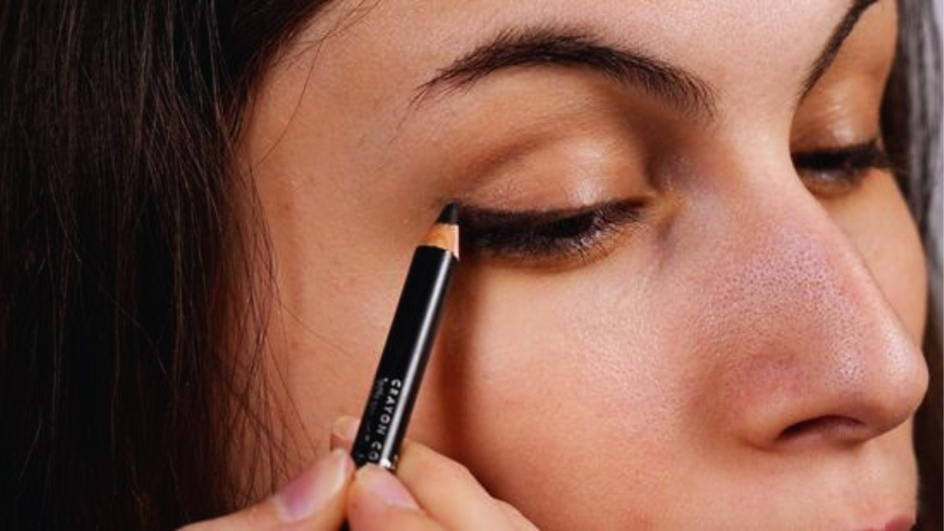 Close-up of a person applying black eyeliner to their upper eyelid, demonstrating a makeup technique. The scene conveys focus and precision.