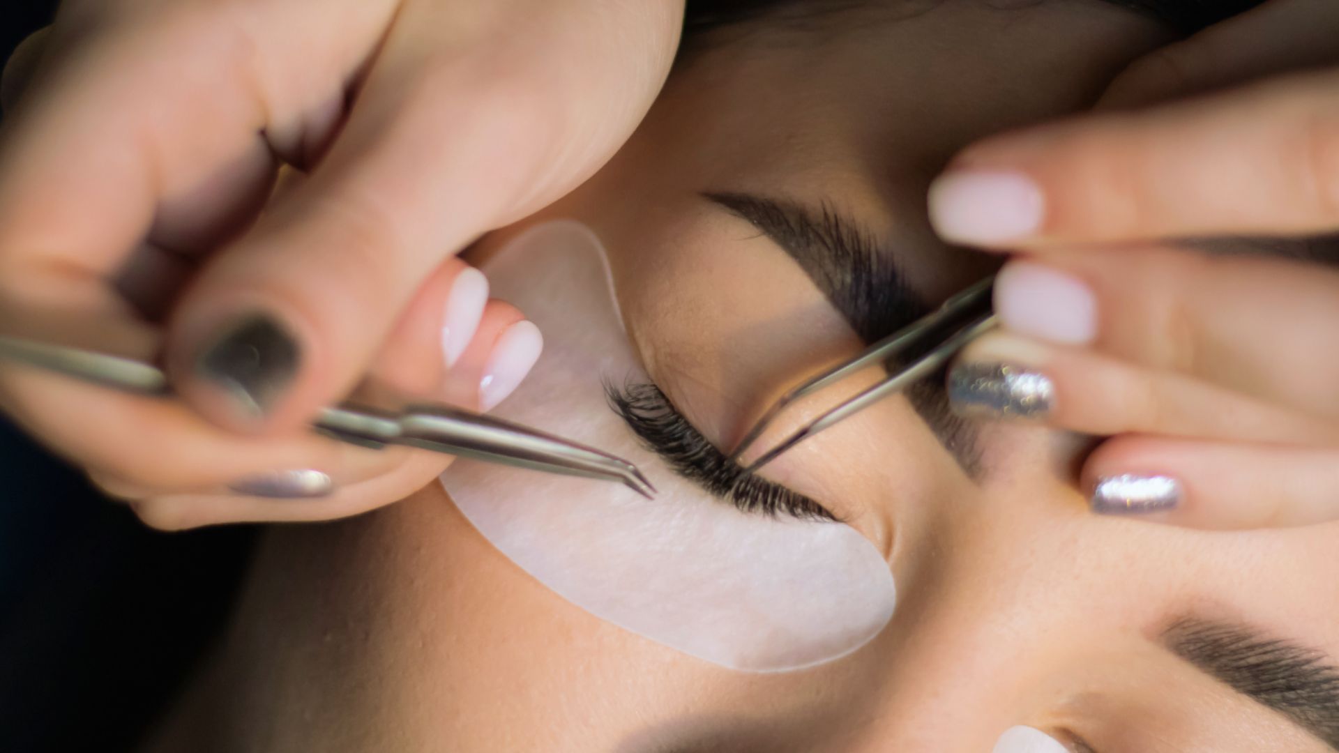Close-up of a person receiving eyelash extensions. Two hands use tweezers to apply lashes, with an eye pad placed under the closed eye. Calm and focused setting.