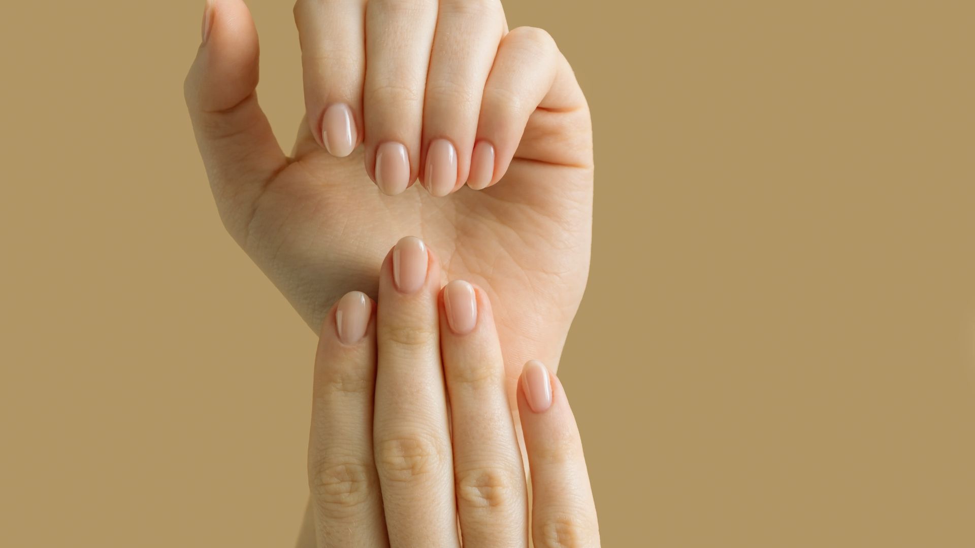 Two hands with neatly manicured, short, nude-colored nails are posed against a beige background, conveying simplicity and elegance.