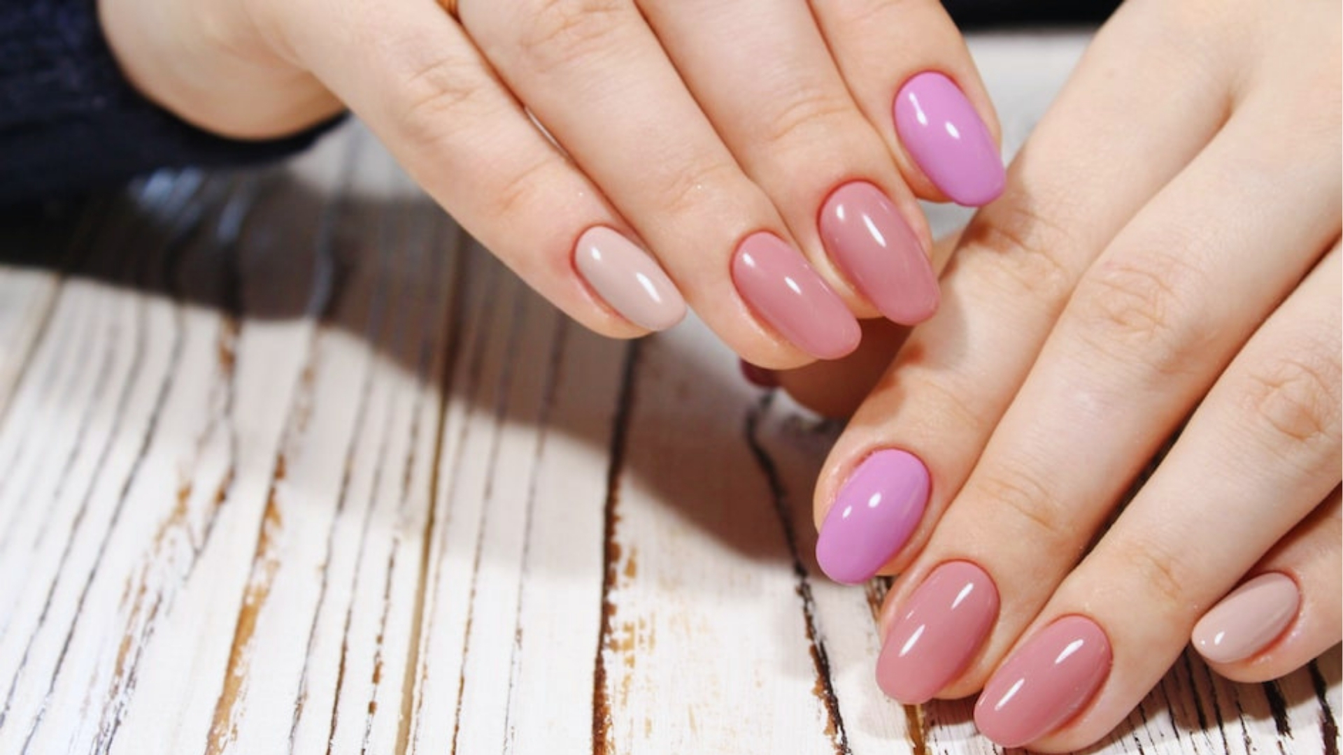 Hands with manicured nails in a gradient of pale pink to vibrant pink polish rest on a textured, whitewashed wooden surface, conveying a stylish and elegant vibe.